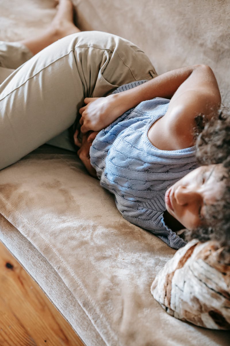 A young woman lying on a couch holding her stomach, indicating pain or discomfort.