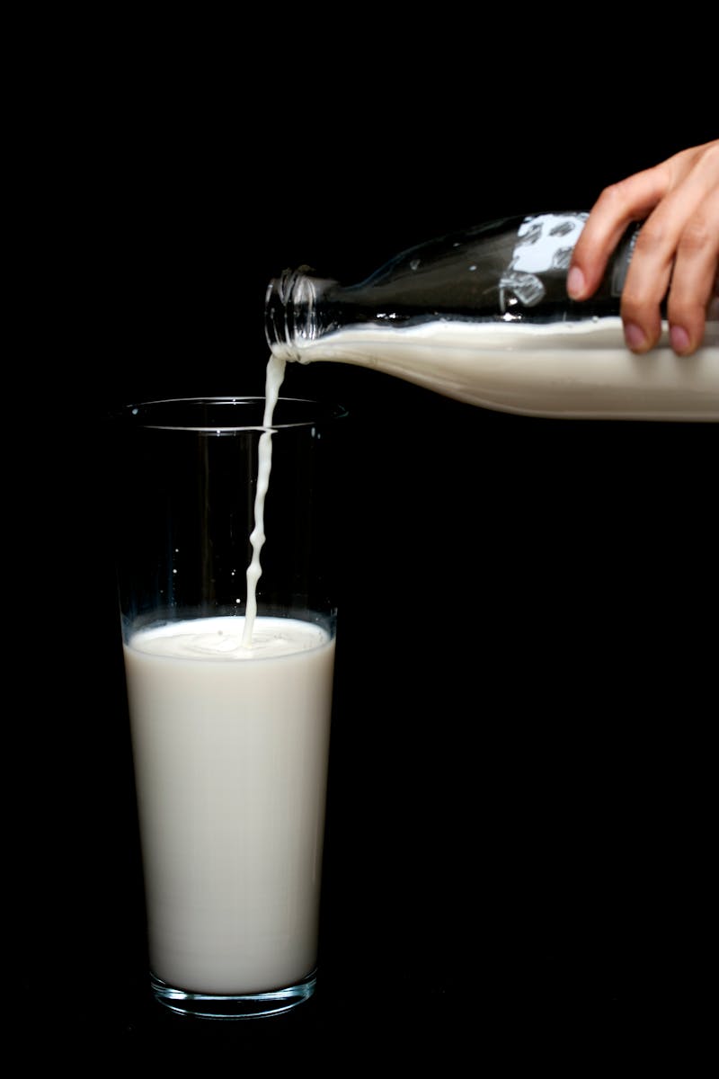 A hand pours fresh milk from a glass bottle into a tall glass with a black backdrop.