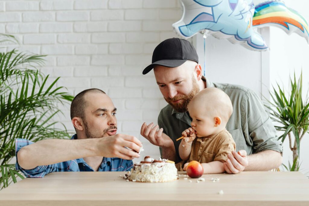Two men celebrating a special moment with their baby, enjoying cake indoors with festive decor.